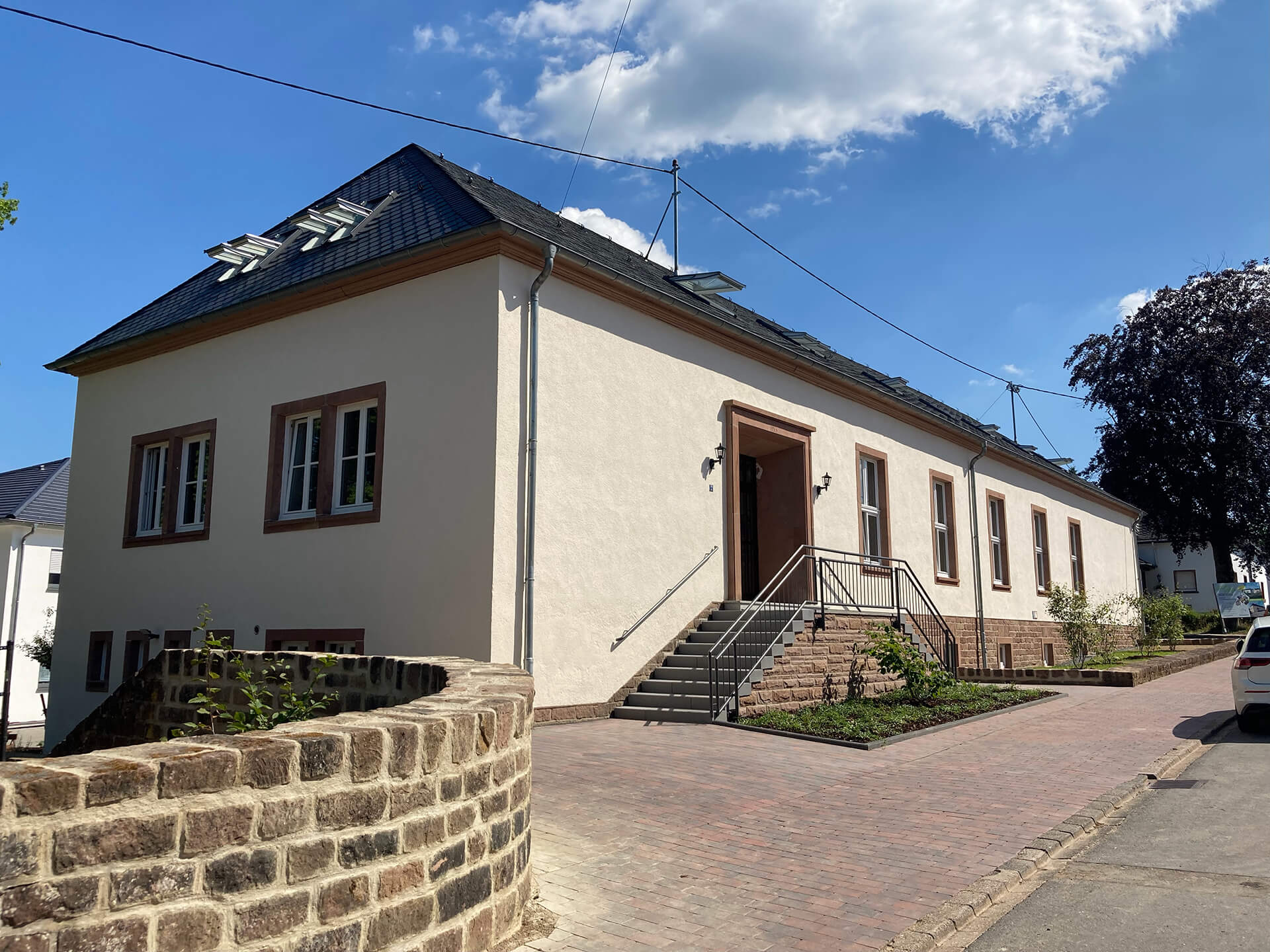 Straßenansicht der umgebauten Alten Schule in Zemmer mit heller Fassade, dunklem Dach und Treppe zum Eingang.