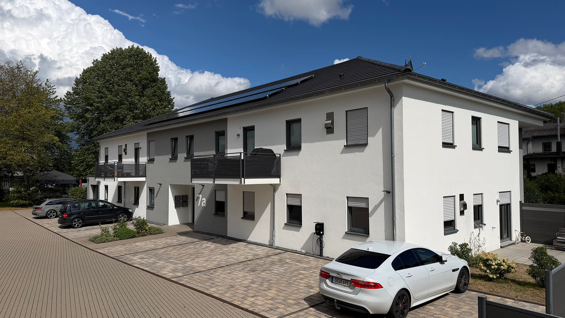 Schräge Ansicht der Eingangs- und Seitenfassade des Mehrfamilienhauses in Zemmer mit dunklen Balkongeländern und Stellplätzen.